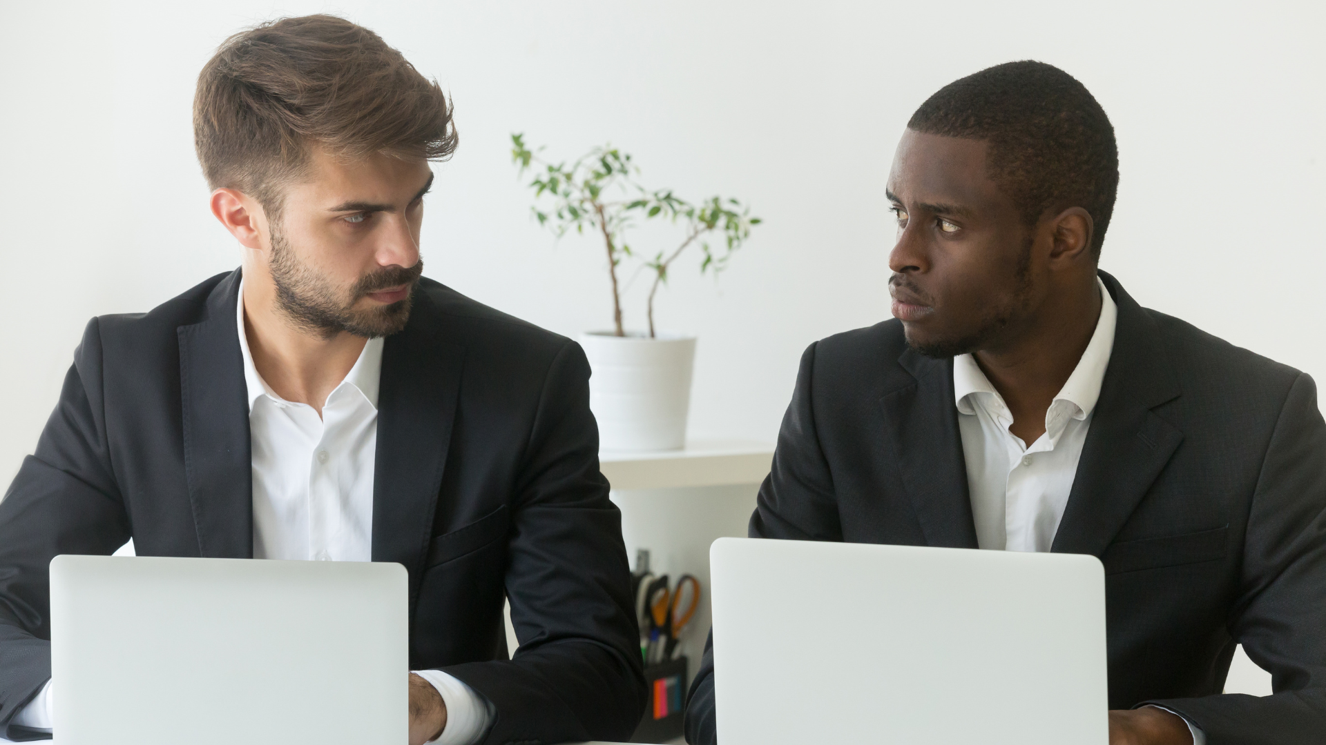 Office rivals sitting beside one another at a desk, looking at each other with an envious stare.